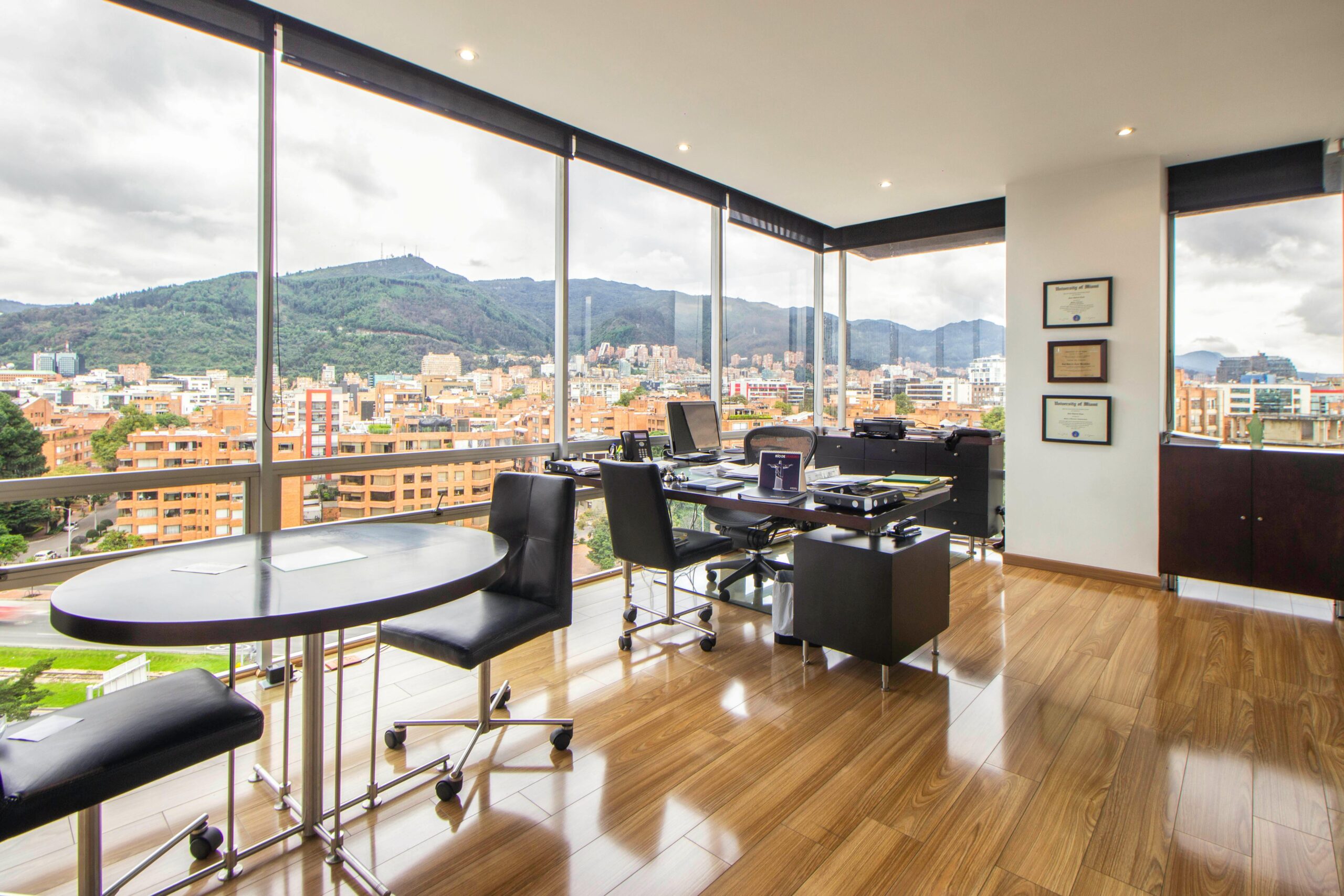 Modern office with floor-to-ceiling windows, city skyline view, wood flooring, and stylish furnishings